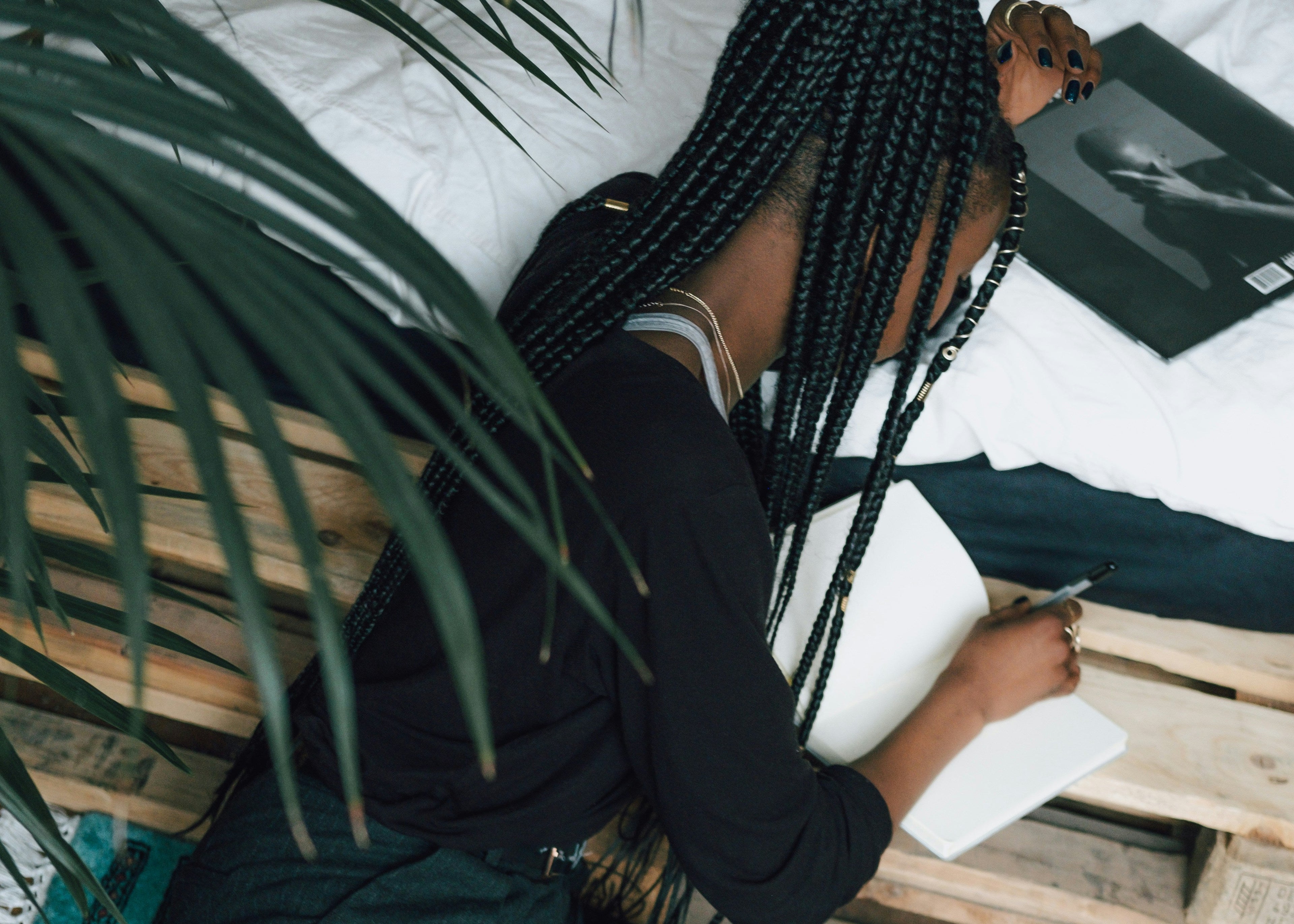 Black woman sitting on floor by bed writing  in notebook with plants overhead and vinyl record