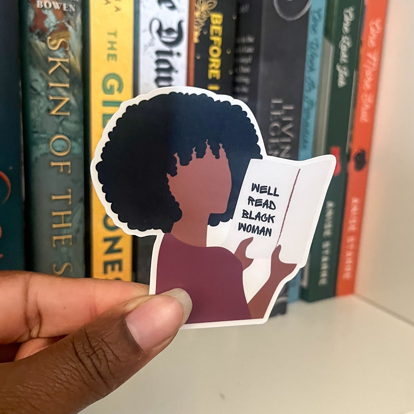Hand holding a sticker of a person with an afro and 'Well Read Black Women' text, with books in the background.