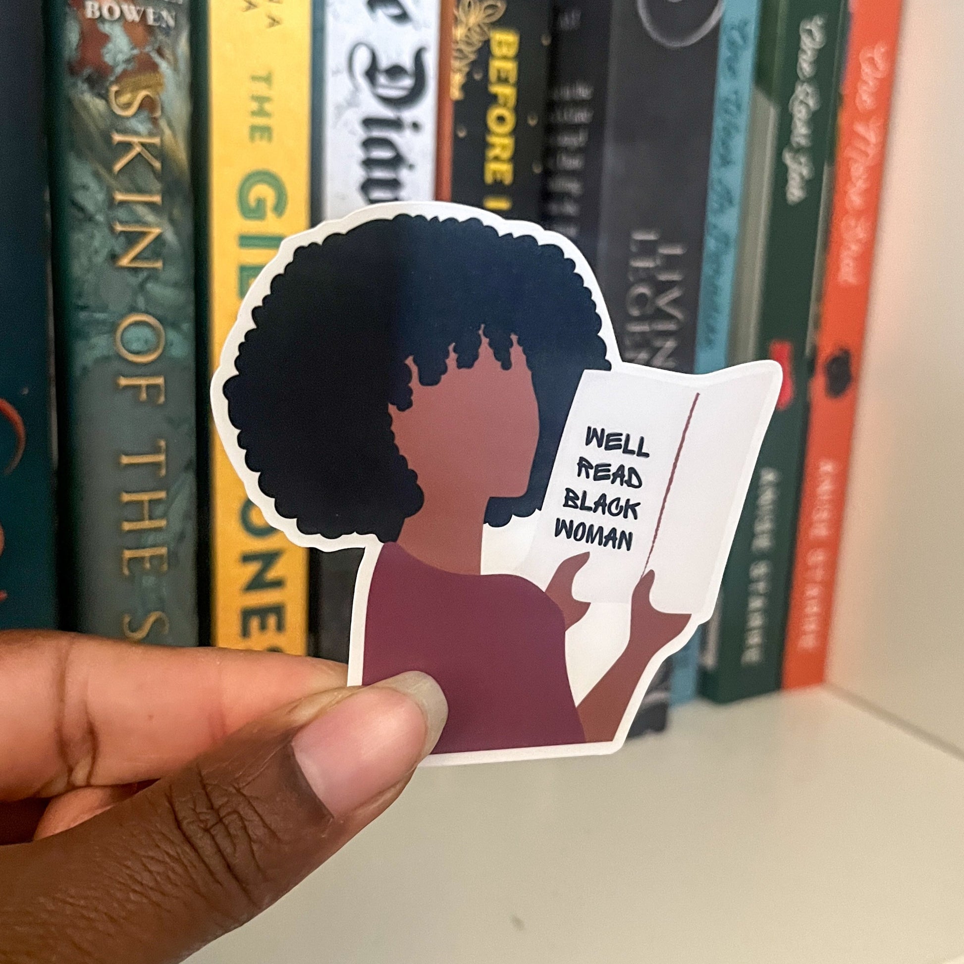 Hand holding a sticker of a person with an afro and 'Well Read Black Women' text, with books in the background.